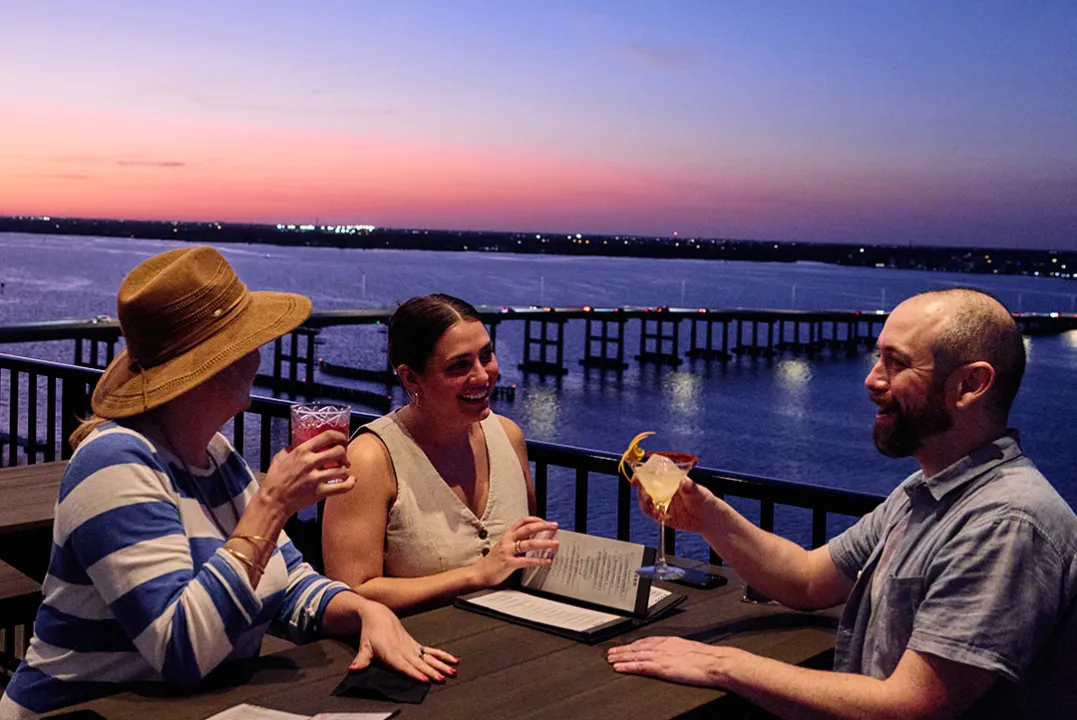 Diners enjoy drinks on a waterfront patio at sunset.