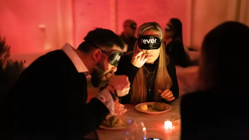 A group of people participate in a blindfolded dining experience in a dimly lit restaurant with a warm, red-toned ambiance. A man and a woman in the foreground wear black blindfolds labeled "Fever" while eating from plates of food. A small candle on the table provides soft lighting, and other blindfolded diners are visible in the background.