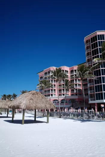 Thatch-roof palapa huts on white sand in front of a pink resort.