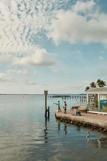 Two people fish from the waterfront at Bat House Park in Matlacha beside a small mural-covered building.