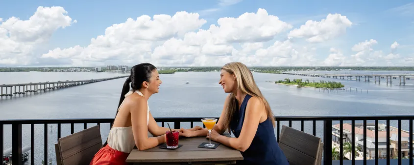 Two women sitting at an outdoor balcony table overlooking a wide river and bridges, smiling and holding cocktails under a bright blue sky with scattered clouds.
