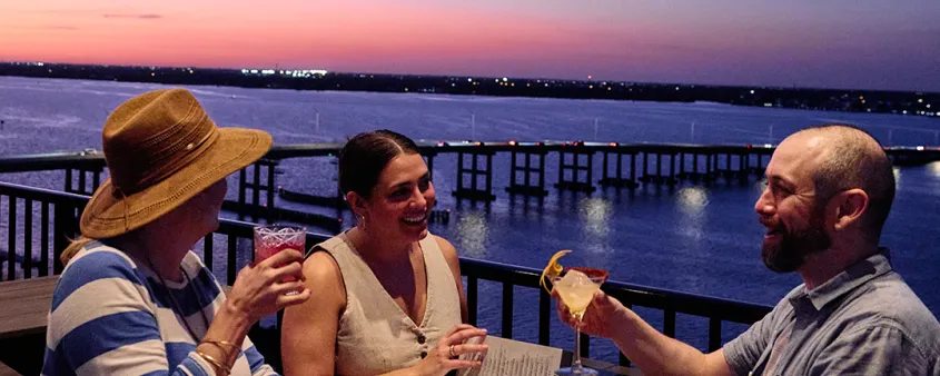 Diners enjoy drinks on a waterfront patio at sunset.