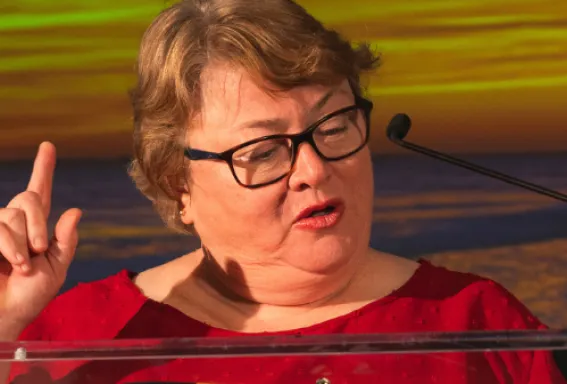 Woman speaking at a podium during a formal event, gesturing while another woman stands beside her smiling against a sunset-themed backdrop.